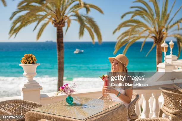 young beautiful smiling blonde woman sitting in the beach restaurant on a table drinking a red cocktail with the turquoise sea  in the background. - blue white summer hat background stock pictures, royalty-free photos & images