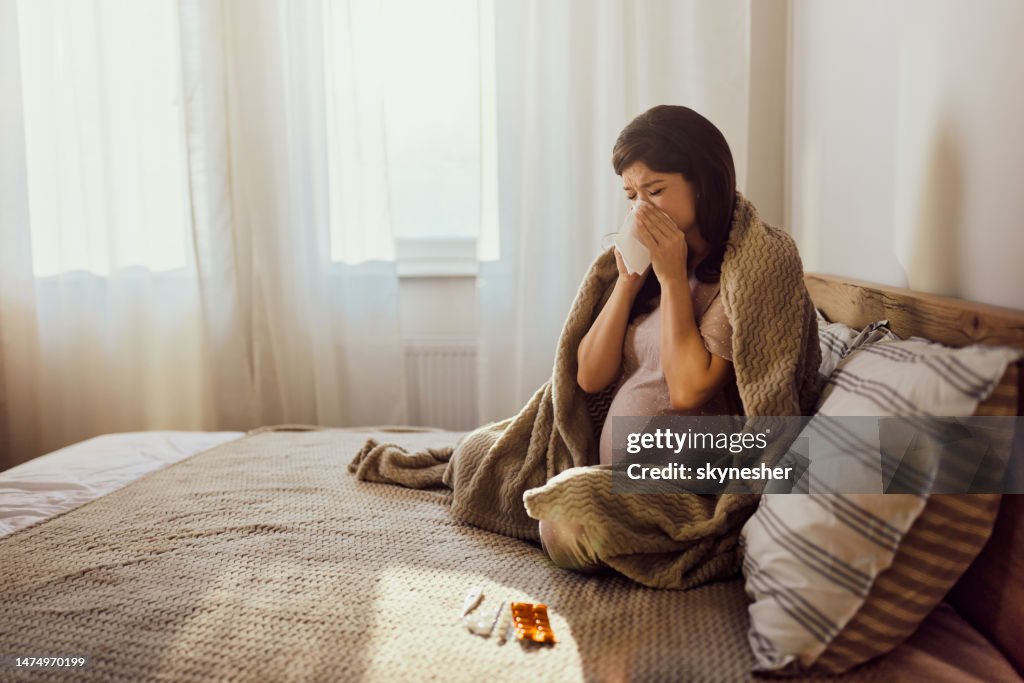 Ill pregnant woman blowing her nose on a bed.