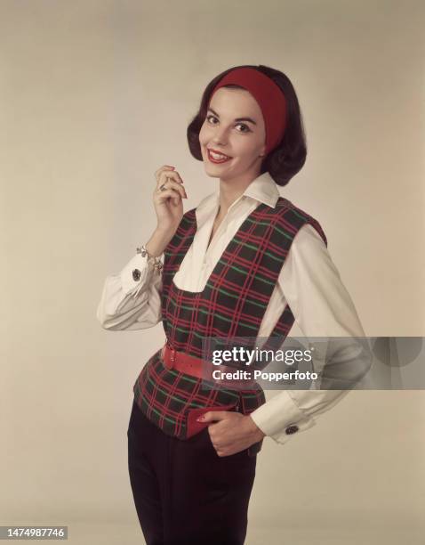 Posed studio portrait of a female fashion model wearing a low necked belted tartan jerkin over a white blouse, a red headband and dark slacks,...