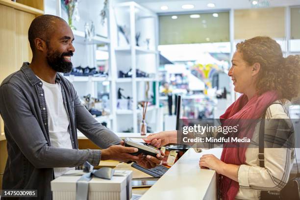 happy female customer placing credit card on credit card reader while friendly male african-american salesperson holds device from behind counter - customer retention stock pictures, royalty-free photos & images