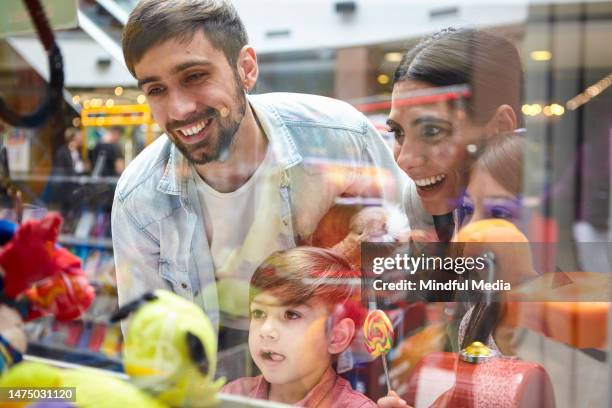 cheerful family standing behind toy grabbing machine's glasses - shopping fun stock pictures, royalty-free photos & images