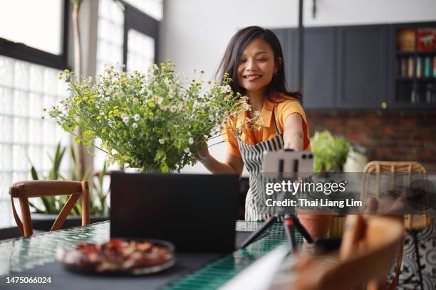 a beautiful asian woman florist is vlogging on her smartphone and laptop, creating an online tutorial on flower bouquet arrangement at home. this scene represents the concept of self-employment, e-learning, online classes, and small business. - social issues stock pictures, royalty-free photos & images
