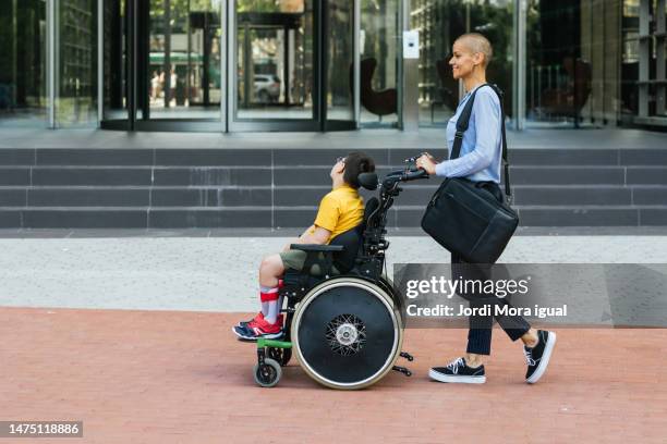 modern mother with laptop bag pushing her son in wheelchair. - quadriplegic stock-fotos und bilder