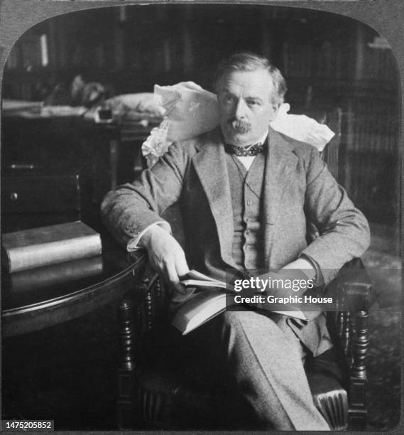 British politician David Lloyd George, Liberal Party MP for Carnarvon Boroughs, wearing a grey three-piece suit with a polka dot bow tie, sits in an...