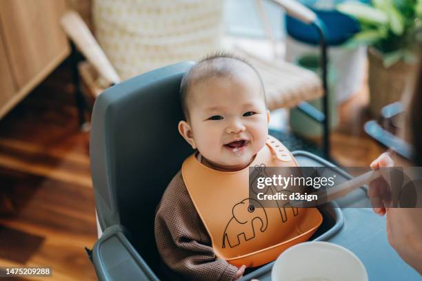 cropped shot of young asian mother feeding her baby daughter for the first time with solid food at home. cute baby girl sitting on high chair smiling at camera. baby milestone, growth and development - よだれ掛け ストックフォトと画像