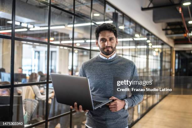 portrait of a mid adult businessman holding laptop in the corridor at office - hispanic businessman stock pictures, royalty-free photos & images