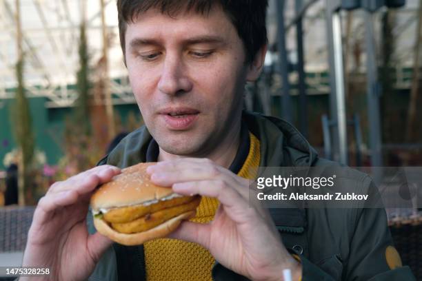 a young man is holding a piece of hamburger in his hands. a guy or a man eats fast food. a hungry skinny guy is eating an appetizing burger. the concept of unhealthy food, diet, overeating, gluttony, dependence on food. fast food restaurant, snack bar. - georgian man stock pictures, royalty-free photos & images