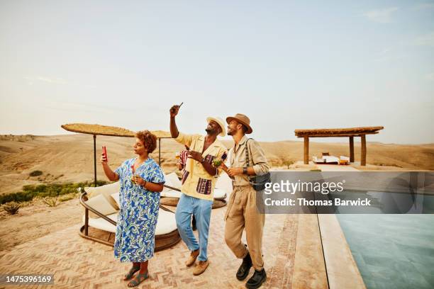 wide shot smiling friends taking selfies by pool at desert camp - desert pool party stock pictures, royalty-free photos & images