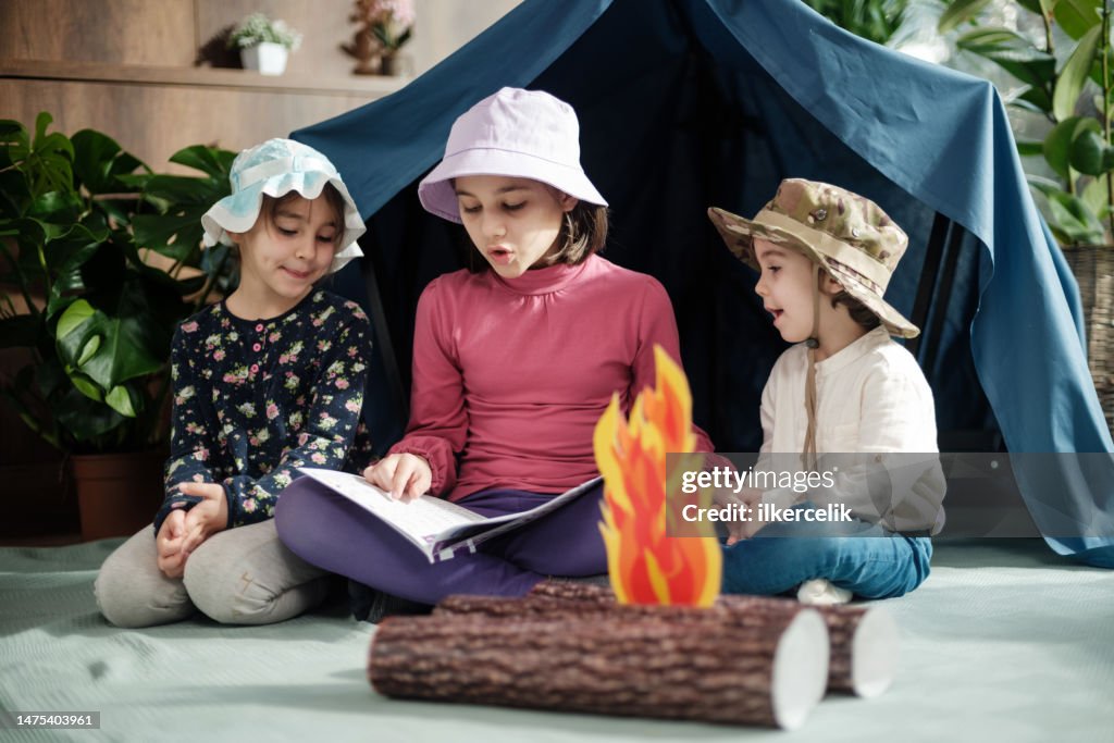 Two child girls and one toddler boy playing camping game and reading book at home, concept for activities for activities at home for children