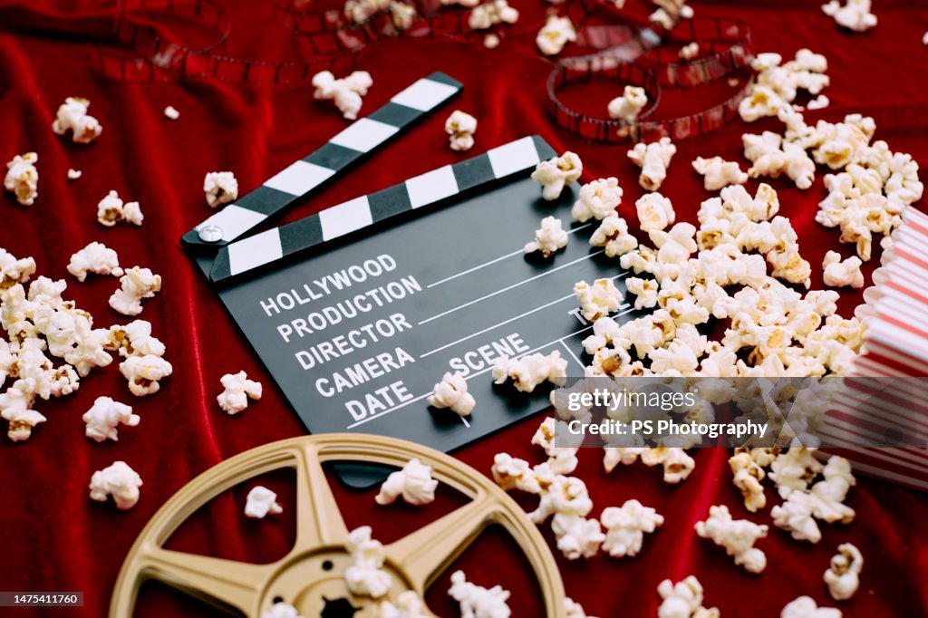 Clapperboard, film reel, film and spilled popcorn on red satin background.