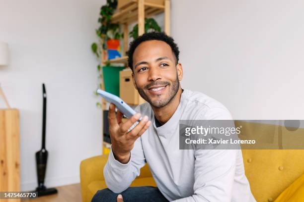 young handsome latin american man using smartphone while sitting on sofa at home. smiling millennial guy sending audio voice message through mobile phone app. trends, social media, technology and domestic lifestyle concept. - mid adult men stock pictures, royalty-free photos & images