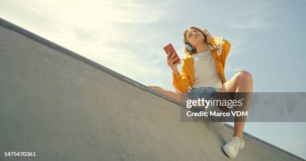 skatepark, headphones and woman on a phone listening to music for calm, gen z  summer holiday on blue sky. young person with streetwear and cellphone, smartphone or 5g audio tech in urban city below - music inspired fashion stock pictures, royalty-free photos & images