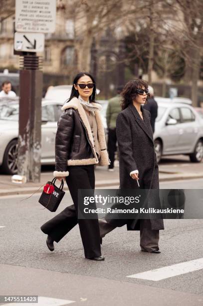 Pornwika Spiecker wears black sunglasses, a white with black print pattern silk scarf, a brown buttoned suit gilet, a black shiny leather with beige...