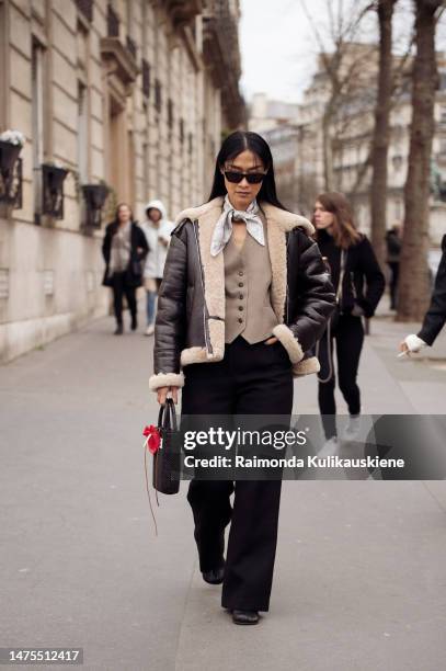 Pornwika Spiecker wears black sunglasses, a white with black print pattern silk scarf, a brown buttoned suit gilet, a black shiny leather with beige...