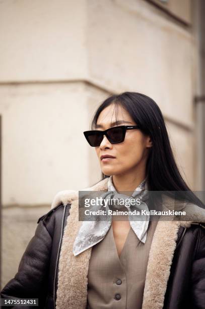 Pornwika Spiecker wears black sunglasses, a white with black print pattern silk scarf, a brown buttoned suit gilet, a black shiny leather with beige...