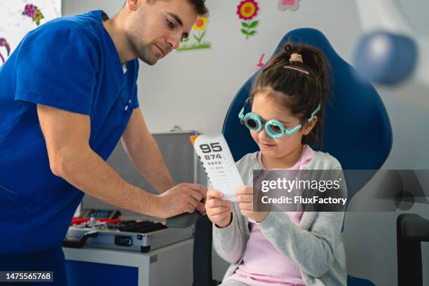 ophthalmologist, examining little girl's eyesight, while using trial frame and different lenses, while she reading snellen chart - ophthalmologist chart stock pictures, royalty-free photos & images