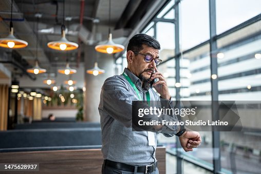 Hombre maduro hablando por teléfono y revisando la hora en su reloj de pulsera en la oficina Hombre maduro hablando por teléfono y revisando la hora en su reloj de pulsera en la oficina