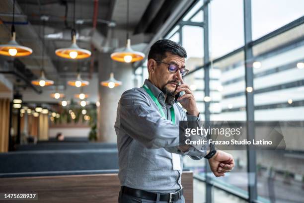 hombre maduro hablando por teléfono y revisando la hora en su reloj de pulsera en la oficina - tiempo fotografías e imágenes de stock