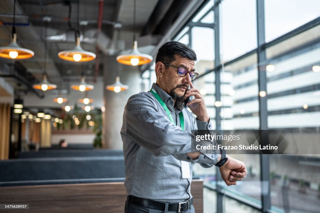 Mature man talking on the phone and checking the time on his wristwatch at office