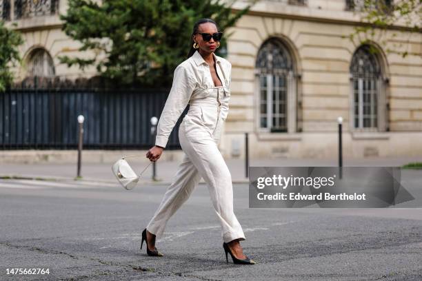 Emilie Joseph wears golden earrings, sunglasses, a white Machou shirt from Jacquemus, a white linen jumpsuit / corset "L'Année 97" by Jacquemus, a...