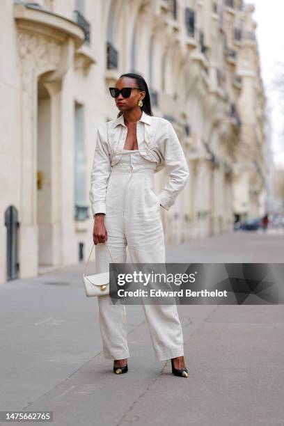 Emilie Joseph wears golden earrings, sunglasses, a white Machou shirt from Jacquemus, a white linen jumpsuit / corset "L'Année 97" by Jacquemus, a...