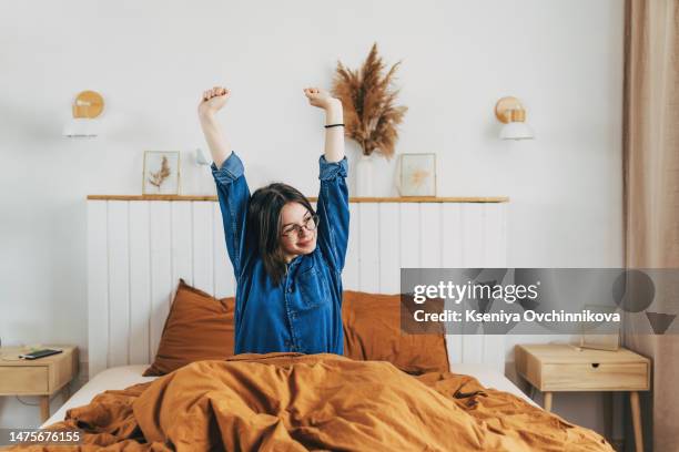 happy blonde-hair woman is sitting on the bed, stretching after sleep, smiling. attractive young female in stylish sportswear warm up before workout, morning training concept. high quality photo - amplified heat stock pictures, royalty-free photos & images