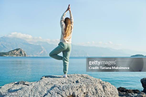 young woman practicing yoga on the stones, beautiful sea background. full length. - tree position stock pictures, royalty-free photos & images