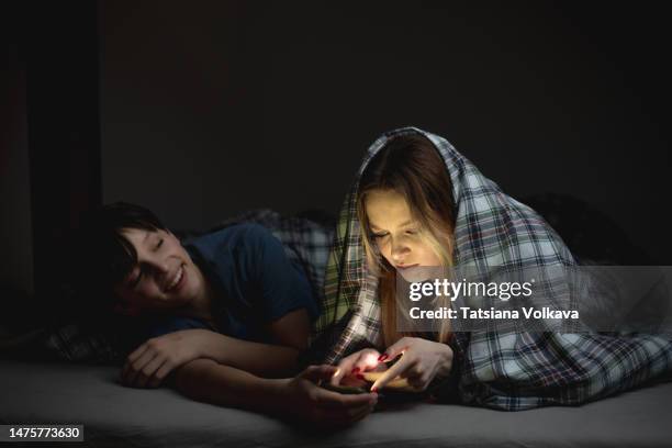 teen boy and teen girl playing games on mobile phones while lying on bed in bedroom under blanket, spending time together hiding from parents on night - annoying brother stock pictures, royalty-free photos & images