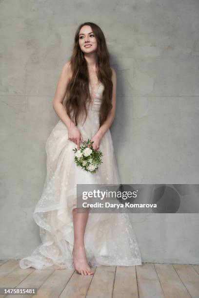 beautiful young woman with long brown hair in wedding dress without shoes and with flowers on grey background with copy space. the happy woman is smiling. the concept of tired feet during the wedding day. - beautiful long legs imagens e fotografias de stock