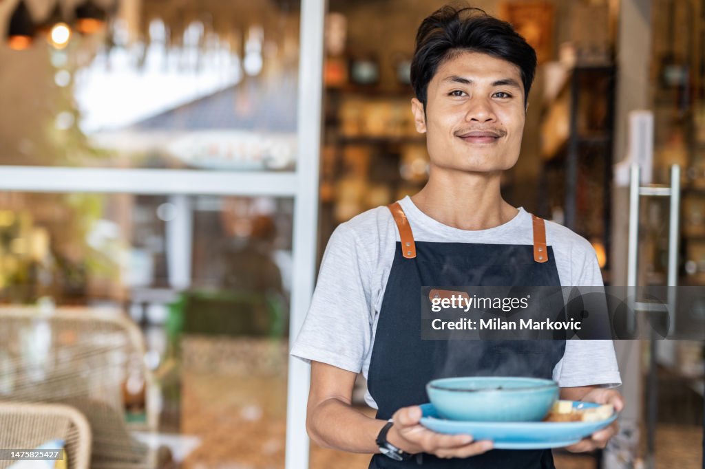 Um garçom indonésio em Bali que serve comida em um restaurante, ele traz sopa quente, um prato tradicional balinês