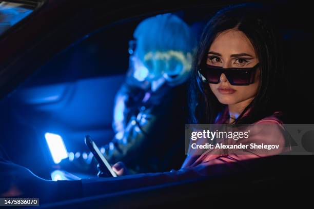 未来的な眼鏡をかけ、車内で携帯電話を使用する2人の女性のポートレート。フィルムノワールスタイルとサイバーパンクのコンセプト写真、ネオンライトの映画写真。青い髪型、人と乗り物 - smartphone noir ストックフォトと画像