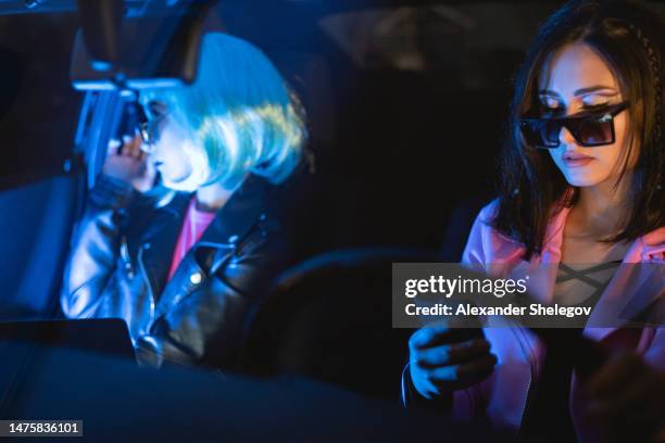 未来的な眼鏡をかけ、車内で携帯電話を使用する2人の女性のポートレート。フィルムノワールスタイルとサイバーパンクのコンセプト写真、ネオンライトの映画写真。青い髪型、人と乗り物 - smartphone noir ストックフォトと画像