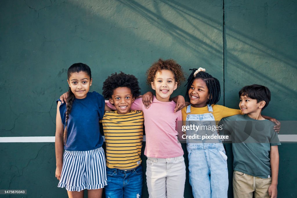 Diversity, friendship and portrait of children on a playground hugging after playing together. Happy, smile and multiracial young kid friends standing, embracing and bonding by a wall on park in city
