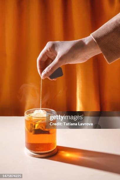 hand making tea with lemons by putting tea bag into transparent glass against orange curtains background with beautiful illuminated shadow.  closeup of natural hot beverage. front view - herbal tea bag stock pictures, royalty-free photos & images