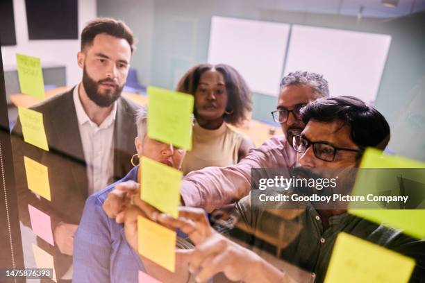 diverse geschäftsleute brainstormen gemeinsam mit klebenotizen auf einer glaswand - team looking at adhesive notes in board room during meeting stock-fotos und bilder