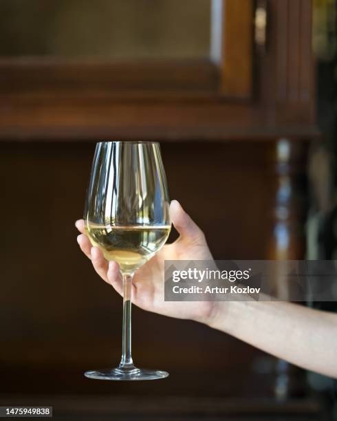 glass of white wine in woman's hand. wine tasting. examining color of wine. grape wine golden color. woman raises her glass. wooden blurred background. side view. copy space. - pinot grigio wine foto e immagini stock