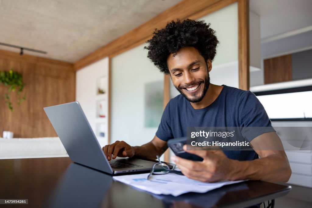 Empreendedor feliz que trabalha em casa usando seu laptop e seu telefone celular