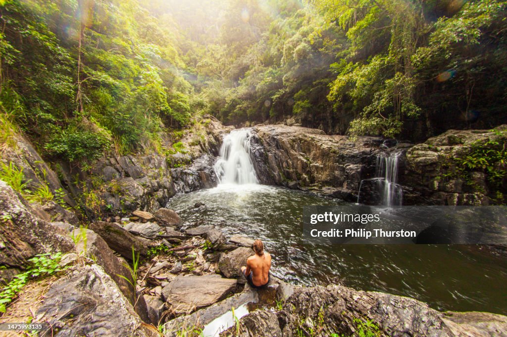Young man sitting by waterfall flowing over rocks into freshwater swimming hole in lush tropical rainforest scene