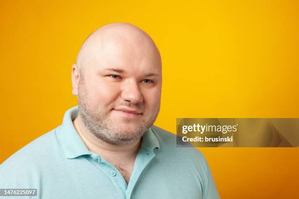 ritratto in studio ravvicinato di uomo bianco adulto medio sovrappeso con testa rasata, che indossa una polo blu, su uno sfondo giallo - corporatura larga foto e immagini stock