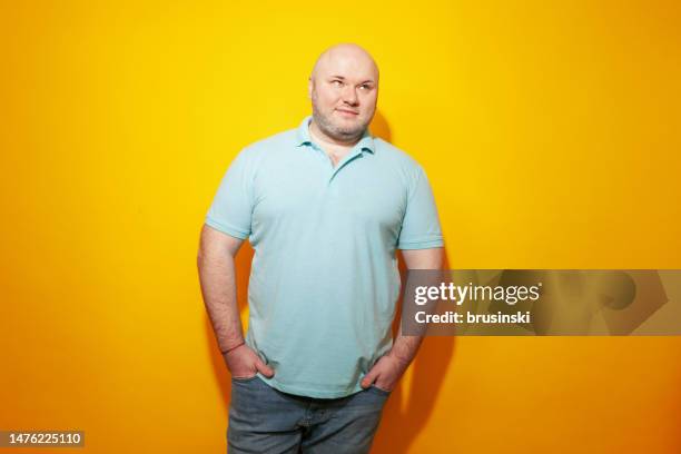 studio portrait of overweight mid adult white man with shaved head, wearing blue polo shirt, against a yellow background - mid adult men stock pictures, royalty-free photos & images