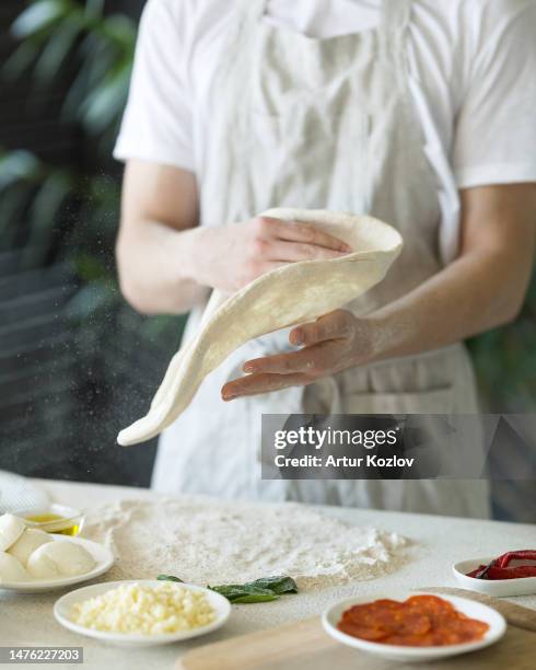 process of making pizza. pizzaiollo in white overalls is kneading and shaping dough. ingredients for pizza: cheese, sauce, pepper, flour on table. basis for pizza. kitchen. front view. soft focus - pizza chef stock pictures, royalty-free photos & images