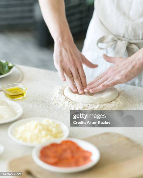 process of making pizza. pizzaiollo in white overalls is kneading and shaping dough. ingredients for pizza: cheese, paperoni, olive oil on table. pizza base. pizzeria kitchen. front view. soft focus - pizza chef stock pictures, royalty-free photos & images