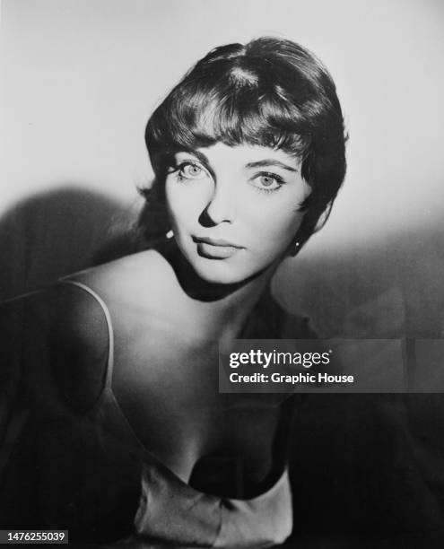 British actress Joan Collins wearing a scooped neck outfit with spaghetti straps, her face illuminated while the rest of the image is cast in...