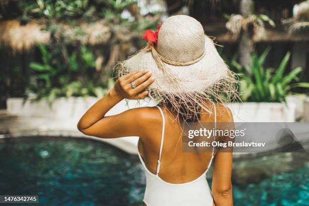 view from behind of young tanned woman in straw hat and swimsuit, swimming pool on background - bali luxury stock pictures, royalty-free photos & images