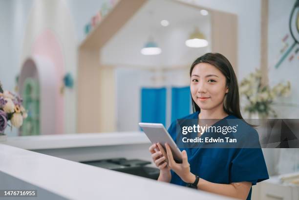 retrato de una enfermera china asiática en el mostrador de la recepcionista del vestíbulo mirando a la cámara sonriendo - recepcionista fotografías e imágenes de stock