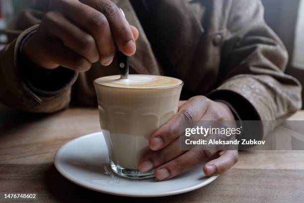 a cup of coffee on the table, in a cafe, cafeteria, coworking or at home. coffee with vegetable milk in the hands of a dark-skinned man. coffee cup. the concept of loneliness, a business breakfast. coffee break. a young businessman drinks coffee. - hand resting on wood stock-fotos und bilder