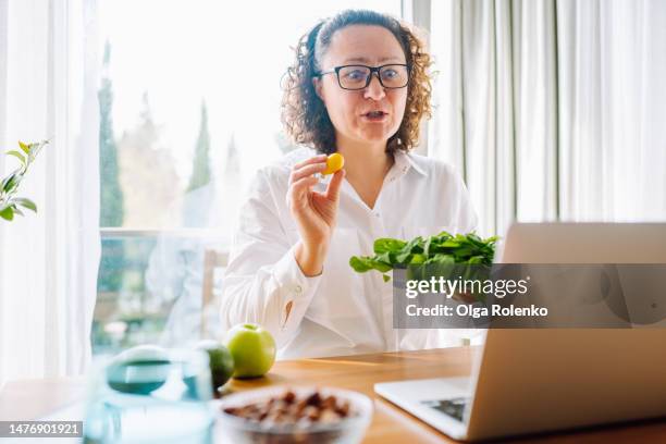 medical nutrition counseling about healthy eating and diet online. woman in lab coat showing organic, vitamin food, streaming online on a laptop in light hospital office - nutrition coach stock pictures, royalty-free photos & images