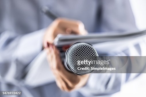 A journalist holds a microphone at a press conference and writes information in a notebook.