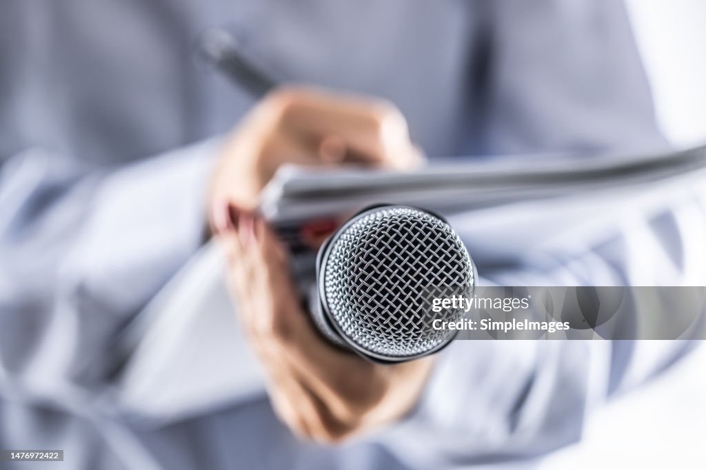 A journalist holds a microphone at a press conference and writes information in a notebook.