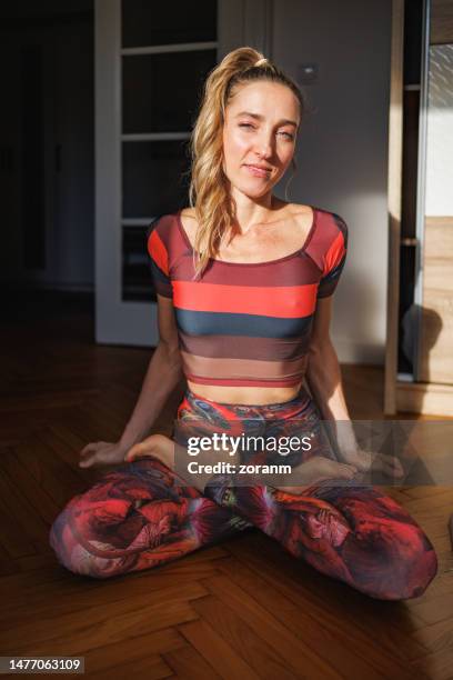 woman doing yoga at home, sitting on hardwood floor in lotus pose and looking at camera - upright position stock pictures, royalty-free photos & images